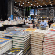 Zu sehen ist eine Messehalle mit vielen Besucher*innen und stapelweise Bücher.