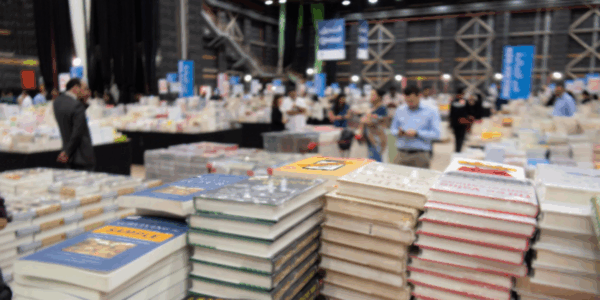 Zu sehen ist eine Messehalle mit vielen Besucher*innen und stapelweise Bücher.