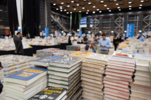 Zu sehen ist eine Messehalle mit vielen Besucher*innen und stapelweise Bücher.